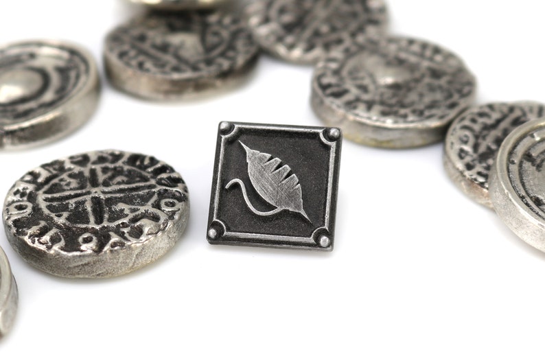May include: A silver metal pin with a black square background and a silver leaf design. The pin is surrounded by several silver coins.