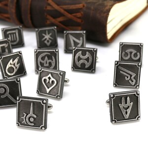 May include: A set of 16 silver cufflinks featuring various symbols, each with a black background and a silver design. The cufflinks are arranged on a white surface.