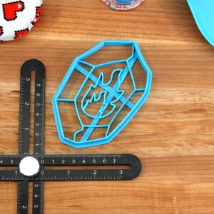 May include: A blue plastic cookie cutter shaped like a flame with a geometric design. The cutter is on a wooden surface.