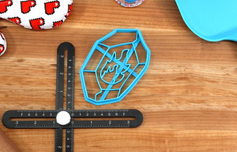 May include: A blue plastic cookie cutter shaped like a flame with a geometric design. The cutter is on a wooden surface.