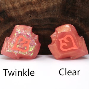 May include: Two pink resin dice with a number 5 on each face. The die on the left has a glittery finish, while the die on the right has a clear finish. The dice are labeled "Twinkle" and "Clear".