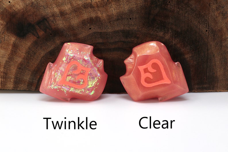 May include: Two pink resin dice with a number 5 on each face. The die on the left has a glittery finish, while the die on the right has a clear finish. The dice are labeled "Twinkle" and "Clear".
