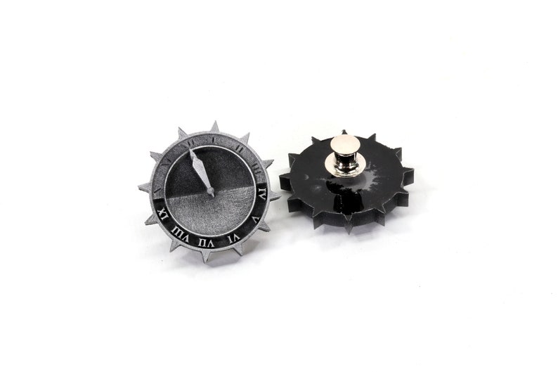 Puede incluir: Dos pines de metal plateados en forma de esferas de reloj con n&uacute;meros romanos. Un pin tiene una esfera negra y el otro tiene una esfera blanca con una aguja negra.