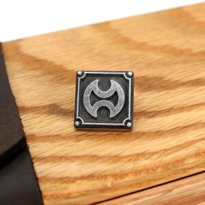 May include: A silver metal square with a stylized design inside. The square has a raised border and four small rivets on each corner. The design is a circle with two half-circles inside, creating a three-dimensional effect.