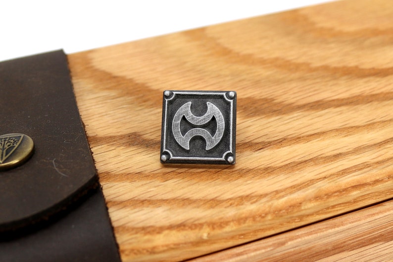 May include: A silver metal square with a stylized design inside. The square has a raised border and four small rivets on each corner. The design is a circle with two half-circles inside, creating a three-dimensional effect.
