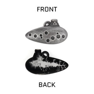 Puede incluir: Un colgante de ocarina plateado con un dise&ntilde;o triangular en la parte superior y una serie de agujeros en la parte delantera. La parte posterior del colgante es negra con un dise&ntilde;o blanco.