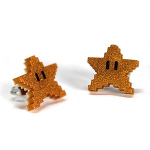 May include: A pair of gold glitter star-shaped earrings with black eyes. The earrings are inspired by the Super Mario Bros. video game.
