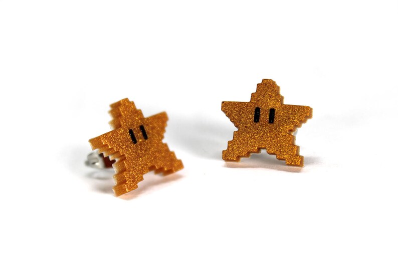May include: A pair of gold glitter star-shaped earrings with black eyes. The earrings are inspired by the Super Mario Bros. video game.