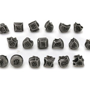 Puede incluir: Un conjunto de 18 pendientes de metal plateado con varios dise&ntilde;os. Cada pendiente presenta un s&iacute;mbolo o dise&ntilde;o diferente, incluyendo formas geom&eacute;tricas, patrones abstractos y caracteres estilizados.