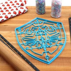 May include: A blue plastic cookie cutter shaped like the Triforce shield from the Legend of Zelda video game series. The cookie cutter is on a wooden surface with a rolling pin, a spatula, and two jars of sprinkles.