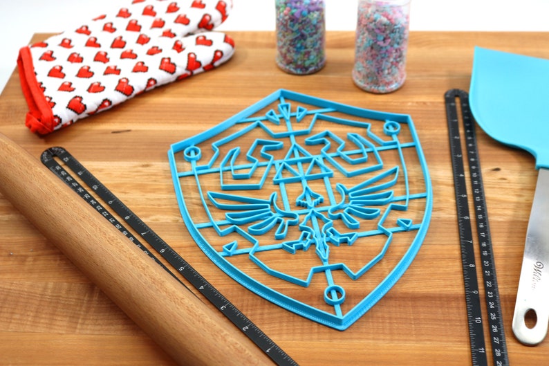May include: A blue plastic cookie cutter shaped like the Triforce shield from the Legend of Zelda video game series. The cookie cutter is on a wooden surface with a rolling pin, a spatula, and two jars of sprinkles.