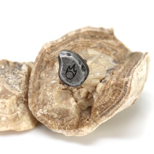 May include: A small, gray, teardrop-shaped pendant with a flame design etched on its surface. The pendant is set on a light brown, textured stone, with a second stone fragment in the background. The background is white.