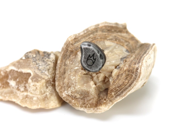 May include: A small, gray, teardrop-shaped pendant with a flame design etched on its surface. The pendant is set on a light brown, textured stone, with a second stone fragment in the background. The background is white.