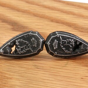 May include: Two black metal shield-shaped buttons with a floral design. The buttons have a raised edge and a small hole in the center.