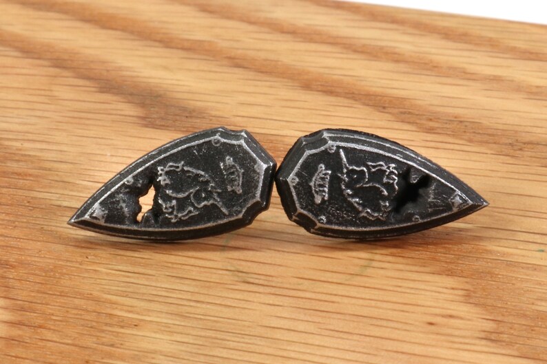 May include: Two black metal shield-shaped buttons with a floral design. The buttons have a raised edge and a small hole in the center.