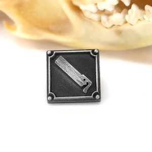 May include: A silver-colored metal pin with a black square background. The pin features a stylized image of a saw blade.