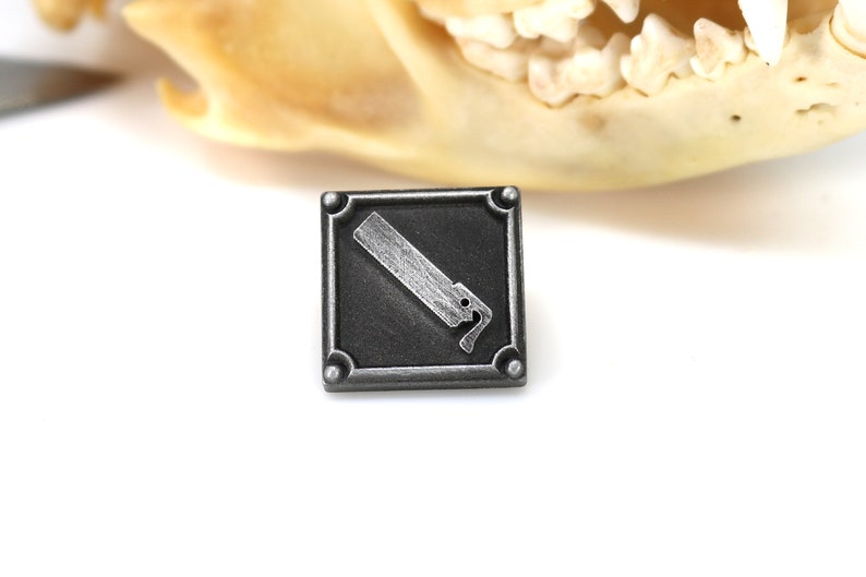 May include: A silver-colored metal pin with a black square background. The pin features a stylized image of a saw blade.