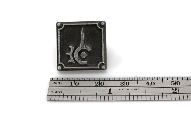 May include: A silver metal pin with a black and silver design. The design is a stylized sword with a circle and spikes around it. The pin is about 2 cm wide.