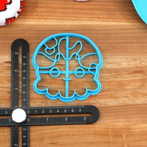 May include: A light blue plastic cookie cutter in the shape of a cartoon character's face. The cutter is on a wooden surface with a black ruler and a white circular object. A red and white patterned fabric is in the background.