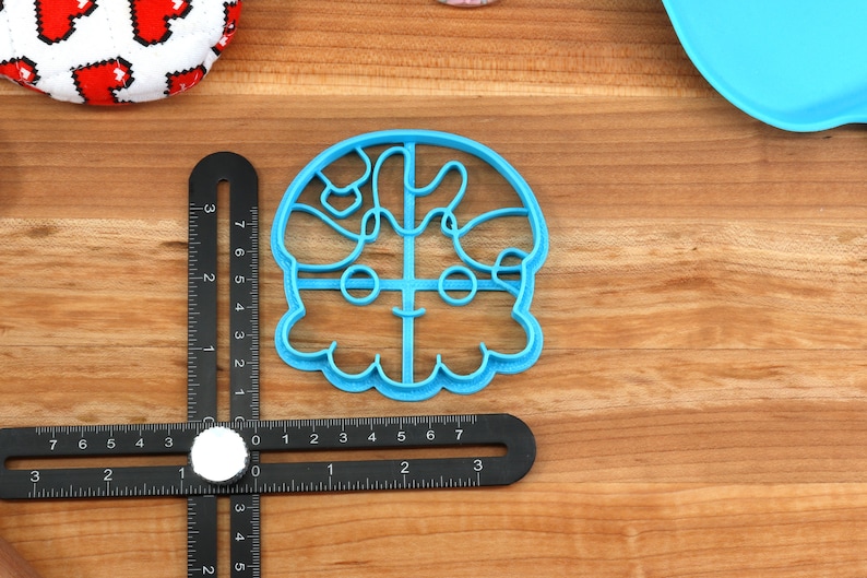 May include: A light blue plastic cookie cutter in the shape of a cartoon character's face. The cutter is on a wooden surface with a black ruler and a white circular object. A red and white patterned fabric is in the background.