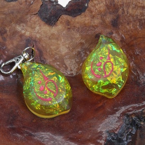 May include: Two iridescent yellow teardrop-shaped keychains with a pink design. The keychains are on a brown wooden surface.