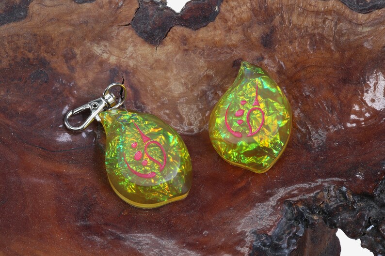 May include: Two iridescent yellow teardrop-shaped keychains with a pink design. The keychains are on a brown wooden surface.