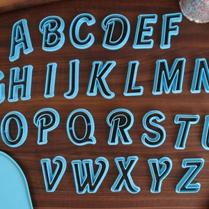 May include: A set of 26 blue and black alphabet cookie cutters on a wooden surface. The cutters are in a cursive font.