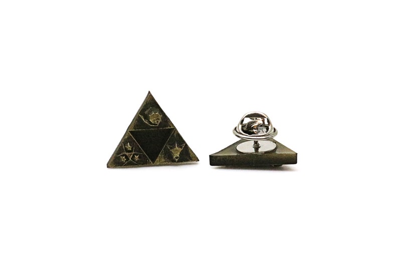 May include: A gold-toned triangular pin with a black and white design. The pin features a stylized image of a triangle with a symbol inside. The back of the pin has a safety clasp.