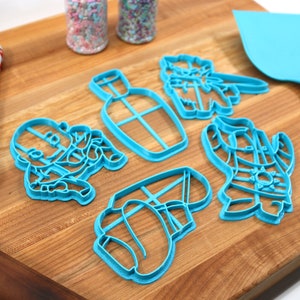 May include: A set of six blue plastic cookie cutters shaped like a bottle, a cactus, a cowboy hat, a cowboy boot, a cowboy, and a cactus. The cutters are on a wooden surface.