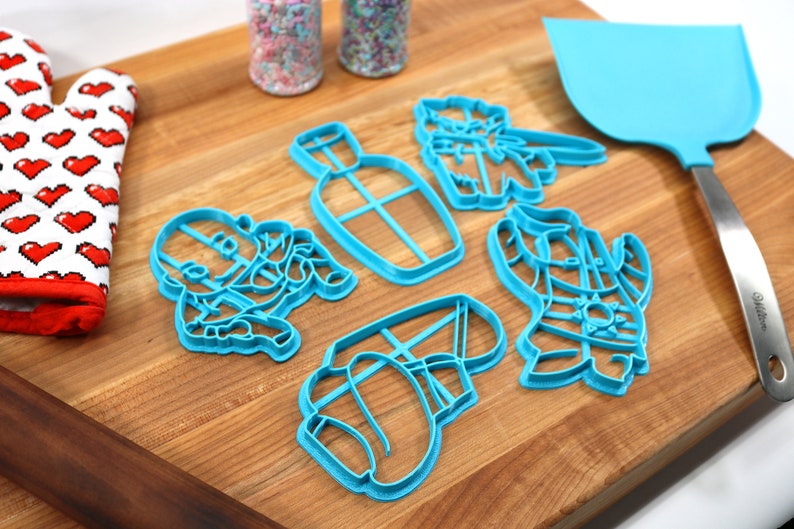 May include: A set of six blue plastic cookie cutters shaped like a bottle, a cactus, a cowboy hat, a cowboy boot, a cowboy, and a cactus. The cutters are on a wooden surface.
