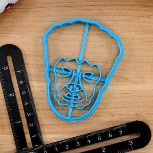 May include: A blue plastic cookie cutter shaped like a person's face with a serious expression.