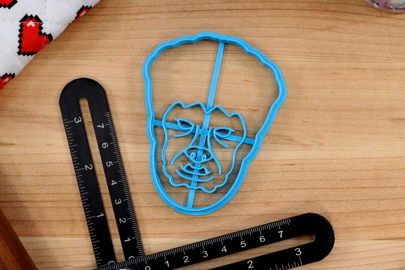 May include: A blue plastic cookie cutter shaped like a person's face with a serious expression.