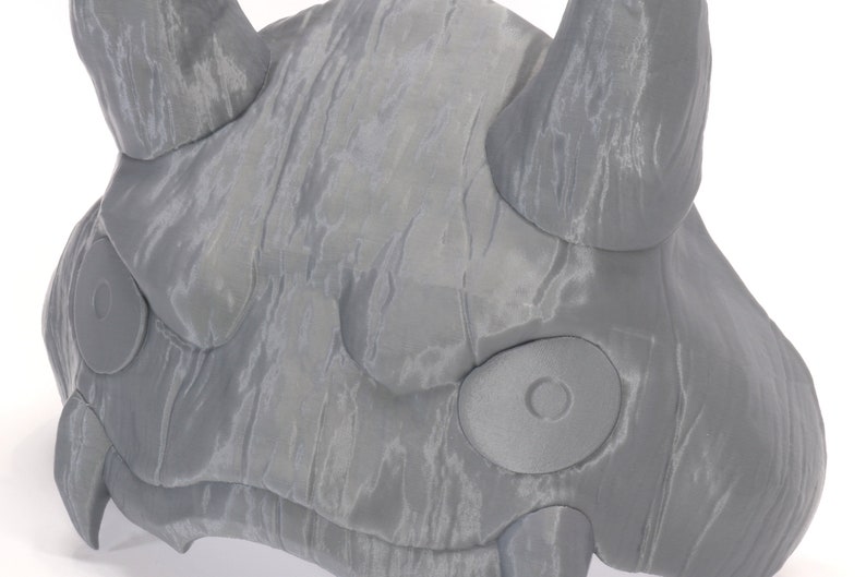 May include: A gray, textured, 3D printed mask with two large, round eyes and two horns. The mask is designed to resemble a mythical creature.