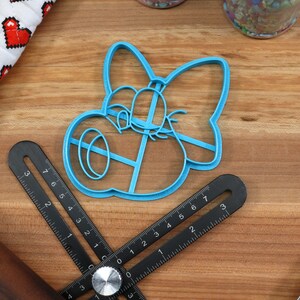 May include: A blue plastic cookie cutter shaped like a cartoon character with a bow. The cutter is on a wooden surface with a ruler and a rolling pin.
