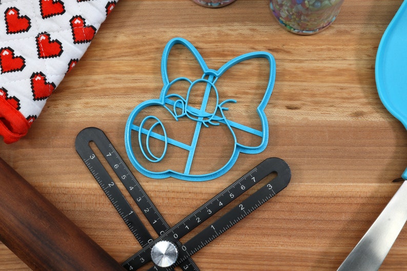 May include: A blue plastic cookie cutter shaped like a cartoon character with a bow. The cutter is on a wooden surface with a ruler and a rolling pin.