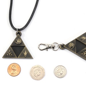 May include: A gold-colored pendant necklace featuring a triangular design with four symbols. The pendant is about the size of a US quarter. The necklace is on a black cord. There is also a matching keychain with a silver clasp.