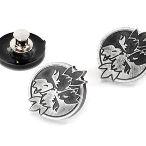 Op de afbeelding: Twee zilverkleurige metalen pinnen met een zwart-wit ontwerp van een gestileerde bloem. De pinnen zijn rond en zijn bevestigd aan een zwarte plastic achterkant.