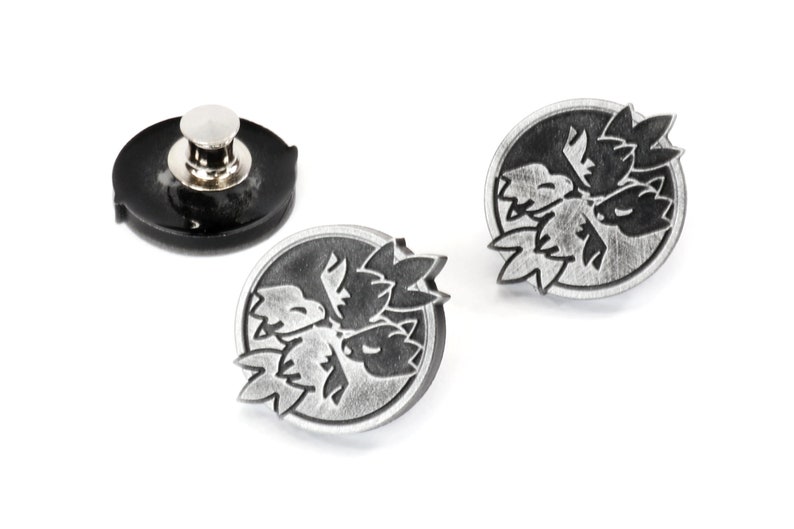 Op de afbeelding: Twee zilverkleurige metalen pinnen met een zwart-wit ontwerp van een gestileerde bloem. De pinnen zijn rond en zijn bevestigd aan een zwarte plastic achterkant.