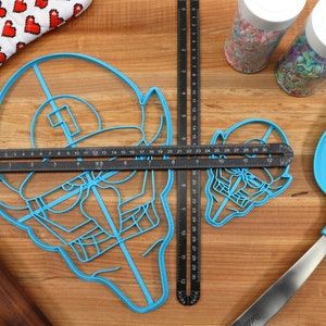 May include: Two blue plastic cookie cutters shaped like a cartoon character with a hat and a mustache. The cutters are on a wooden surface with a ruler, a rolling pin, and a spatula.