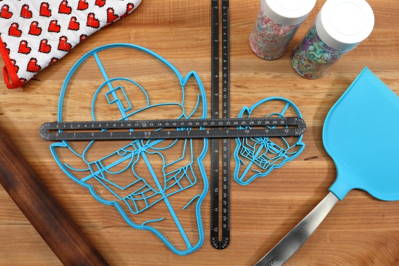 May include: Two blue plastic cookie cutters shaped like a cartoon character with a hat and a mustache. The cutters are on a wooden surface with a ruler, a rolling pin, and a spatula.