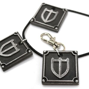 May include: A black leather necklace with three silver square pendants featuring a shield design. The necklace has a silver lobster clasp.