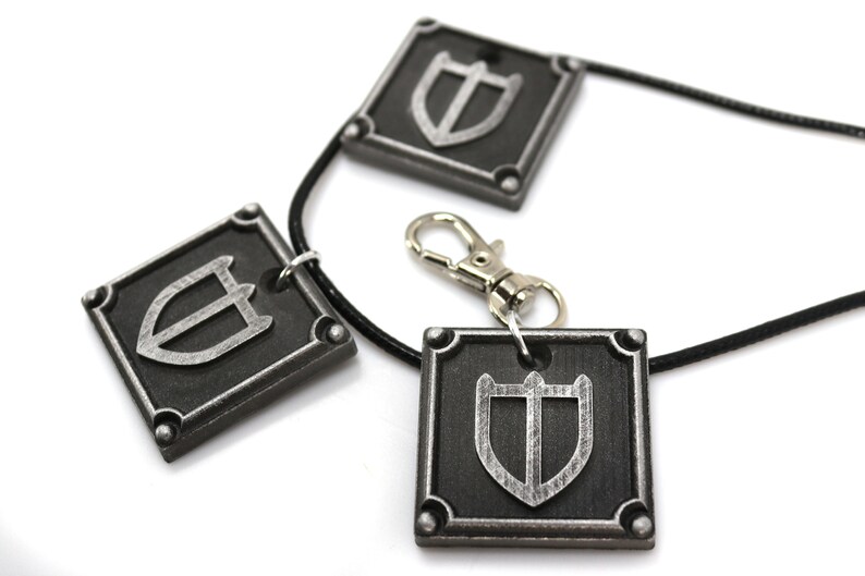 May include: A black leather necklace with three silver square pendants featuring a shield design. The necklace has a silver lobster clasp.
