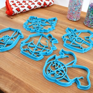 May include: Five blue plastic cookie cutters shaped like different cartoon characters. The cutters are on a wooden surface.