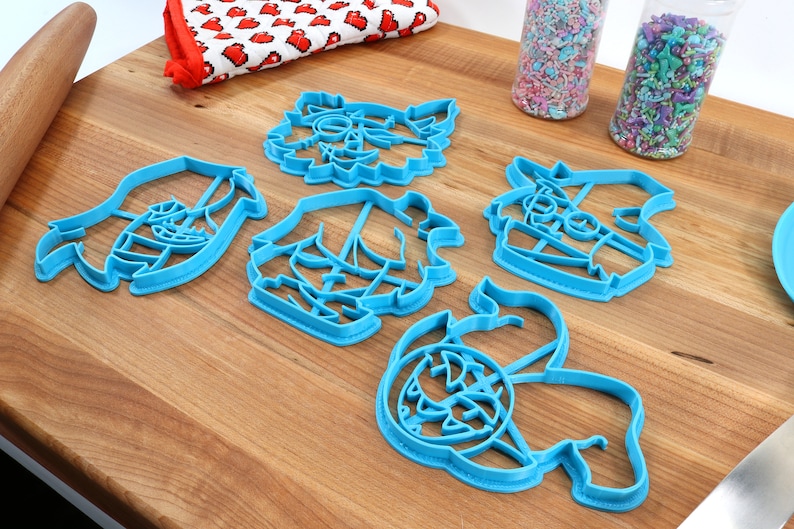 May include: Five blue plastic cookie cutters shaped like different cartoon characters. The cutters are on a wooden surface.