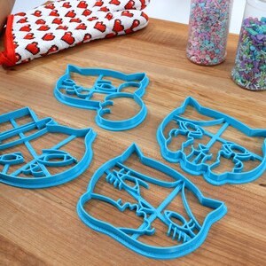 May include: Four blue plastic cookie cutters shaped like cat faces. The cutters are on a wooden surface with sprinkles in jars in the background.