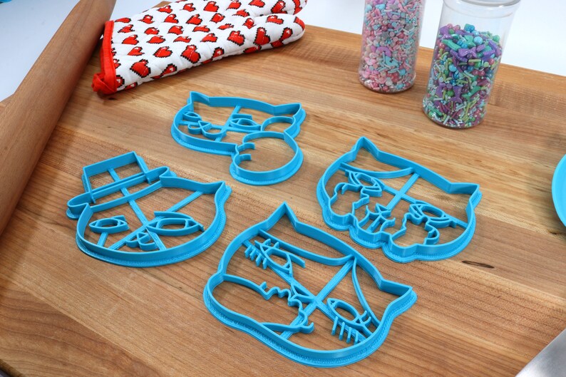 May include: Four blue plastic cookie cutters shaped like cat faces. The cutters are on a wooden surface with sprinkles in jars in the background.