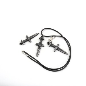 May include: Three silver-toned dagger pendants, one on a black cord necklace. The daggers have intricate details and a pointed tip.