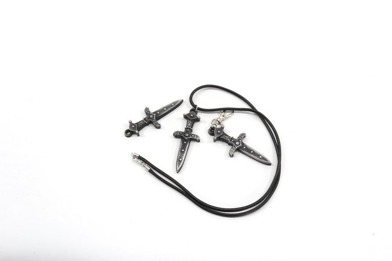 May include: Three silver-toned dagger pendants, one on a black cord necklace. The daggers have intricate details and a pointed tip.