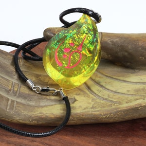 May include: A yellow teardrop-shaped pendant necklace with a pink symbol inside. The necklace is made of black cord and has a silver clasp.