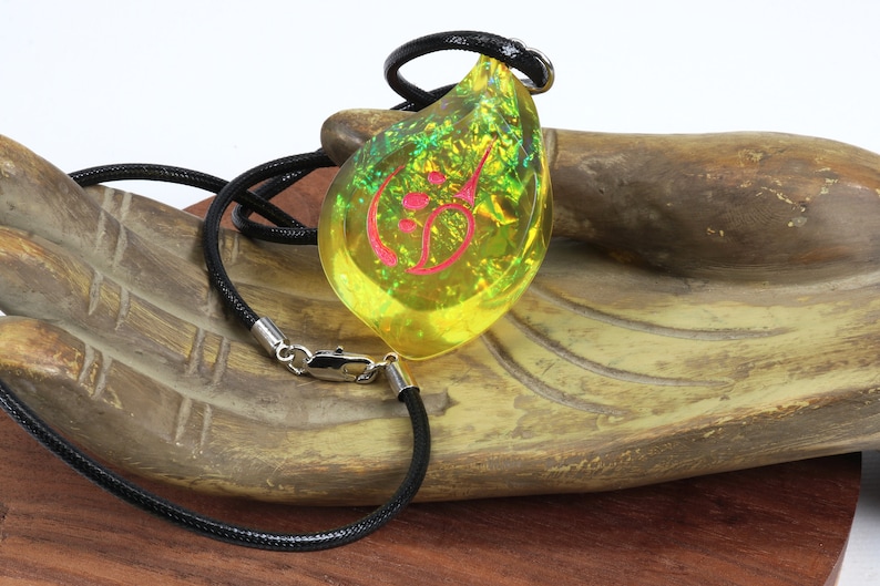 May include: A yellow teardrop-shaped pendant necklace with a pink symbol inside. The necklace is made of black cord and has a silver clasp.